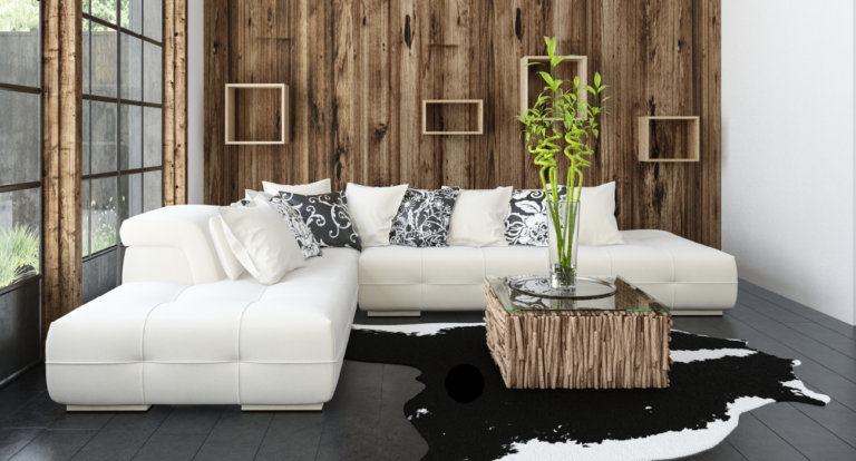 Modern living room with white L-shaped sofa, black and white cushions, wood-paneled wall, rustic coffee table, green plant, large window, and cowhide rug.