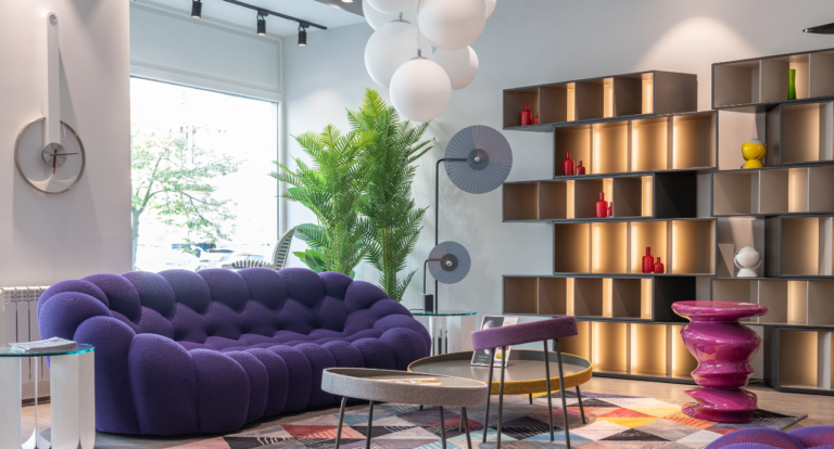 Modern living room with a plush, purple sofa, geometric wall shelves with decorative items, colorful furniture, lush plant, and circular ceiling lights.